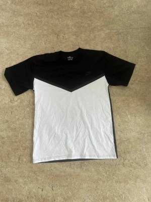 Svartvit t-shirt från Nike - Snygg t-shirt från Nike med coolt svart och vitt blockmönster i V-form framtill. Klassisk rund hals och korta ärmar. Materialet är mjuk bomull som känns skönt mot huden. Perfekt för dig som gillar sportig och stilren streetwear.
