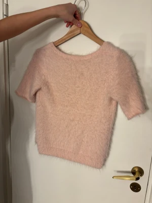 Ljusrosa fluffig stickad kofta - Supermjuk och fluffig ljusrosa stickad kofta med korta ärmar och rund hals. Koftan har en croppad passform och är perfekt för dig som gillar mysiga och trendiga plagg. Materialet är stickat och har en lätt pälsig känsla som gör den extra cozy.