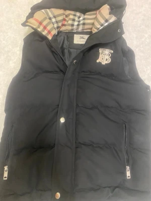 Svart dunväst från Burberry - Säljer en svart dunväst från Burberry med klassiskt rutigt foder i beige, rött och svart. Västen har huva, två fickor med dragkedja och ett broderat Burberry-monogram på bröstet. Perfekt för lager-på-lager och snygg till streetwear.