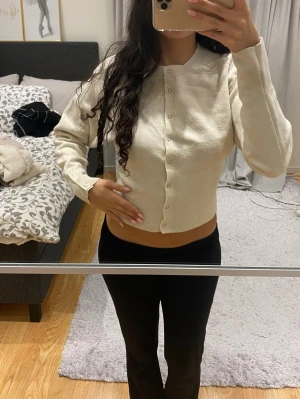 Beige kofta med knappar - Säljer en beige, croppad kofta med rund hals och långa ärmar. Koftan har knappar framtill och även dekorativa knappar vid ärmsluten. Den är stickad och har en clean, enkel look som passar perfekt till jeans eller kjol.