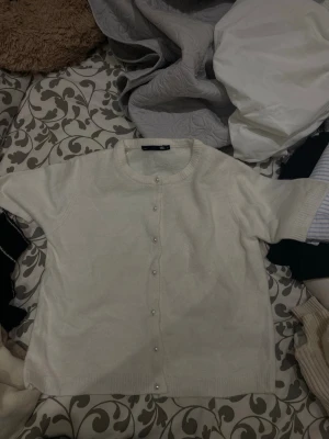 Vit kortärmad kofta med pärlknappar - Säljer en vit kortärmad kofta med rund hals och pärlknappar framtill. Koftan är stickad och har ribbade kanter vid ärmar och nederkant. Perfekt för lager-på-lager och enkel att matcha med olika outfits.