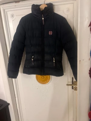 Svart pufferjacka med dragkedjor - Svart pufferjacka från spooks med hög krage och två snedställda fickor med dragkedja. Jackan har quiltad design och ett broderat märke på bröstet. Perfekt för kalla dagar och har en snygg, modern look.