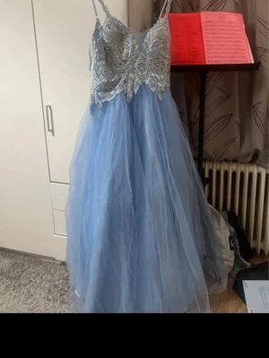 Blå långklänning med glittrig spets - Säljer en blå långklänning med tunna axelband och glittrig spets upptill. Klänningen har ett vackert broderat mönster med paljetter på livet och en voluminös kjol i tyll. Dragkedja i ryggen för enkel påklädning. Perfekt för bal eller fest.