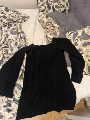 Svart ribbad stickad tröja - Säljer en svart långärmad stickad klänning med ribbad struktur. Klänningen har en rund halsringning och är lite längre i modellen, perfekt att bära över leggings .Materialet är mjukt och ger en skön känsla under kyliga dagar.