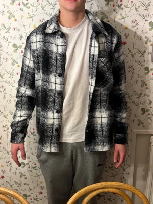 Svartvit rutig overshirt - Säljer en svartvit overshirt med rutigt mönster och klassisk krage. Jackan har knappar framtill och en bröstficka. Perfekt att slänga på över en t-shirt för en chill look. Långärmad och loose passform. 