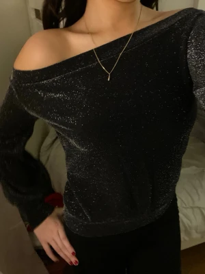 Glittrig svart offshoulder topp - Säljer en svart glittrig topp med offshoulder-design och långa ärmar. Tyget har en skimrande effekt som fångar ljuset och ger en snygg look. Perfekt för dig som vill sticka ut lite extra. Modellen är något loose och toppen är mjuk och bekväm.