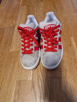 Adidas Campus sneakers beige och röd - Säljer ett par Adidas Campus sneakers i beige mocka med röda detaljer och snörning. Skorna har de klassiska tre ränderna i rött på sidorna och en vit sula med gummisula undertill. Perfekt för dig som gillar retrostil och vill sticka ut med färg.