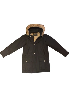 Svart parkajacka från Woolrich med päls - Parkajacka från Woolrich i svart med beige quiltat foder och huva med snygg pälsdetalj. Jackan har stora fickor med knappar och dragkedja framtill. Perfekt för kalla dagar och har en klassisk, stilren look. Passar M/L