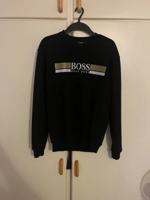 Svart sweatshirt från Hugo Boss - Svart sweatshirt från Hugo Boss med tryckt logga och dekorativa guld- och silverfärgade ränder på bröstet. Tröjan har rund hals och långa ärmar. Materialet är 95% bomull och 5% elastan vilket gör den mjuk och skön att bära.