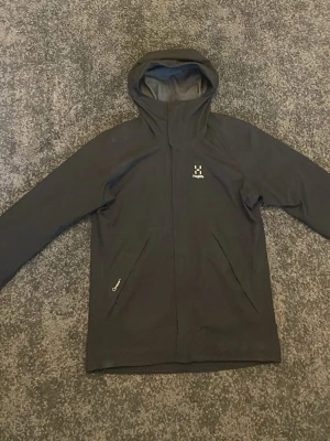 Svart vindjacka från Haglöfs med huva - Svart vindjacka från Haglöfs med snygg minimalistisk design och praktisk huva. Jackan har dragkedja framtill, två snedställda fickor och ett silverfärgat meshfoder på insidan. Perfekt för blåsiga dagar och outdoor-aktiviteter.