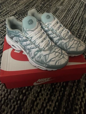 Nike Air Max Plus blå och vit - Nike Air Max Plus sneakers i ljusblått och vitt med mönstrade detaljer och synliga Air Max-enheter i sulan. Skorna har snörning, rund tå och platt sula. Ovandelen är i syntetmaterial med ett unikt blått mönster och vita accenter. Perfekt för dig som gillar statement sneakers.