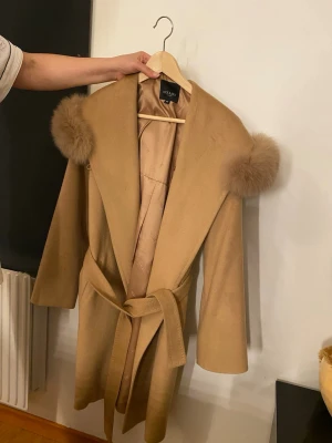 Beige kappa med pälsdetaljer från Hollies - Snygg beige kappa från Hollies med pälsdetaljer på axlarna och ett matchande bälte i midjan. Kappan har en klassisk, rak form och är tillverkad i ullblandning med slät, glansig insida. Perfekt för dig som vill ha en stilren och trendig look.