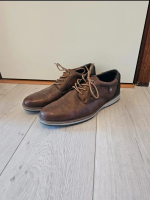 Bruna skinnloafers från Tom Tailor - Snygga bruna loafers i skinn från Tom Tailor med snörning och detaljer i textil på sidorna. Skorna har en platt sula med kontrastfärg och rund tå, vilket ger en stilren och modern look. Perfekta för dig som vill ha en klassisk men ändå trendig sko.