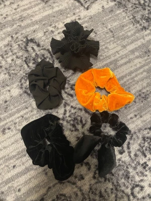 Svart och orange scrunchie-set - Säljer ett set med fem scrunchies i olika stilar och material. Fyra är svarta, varav två i mjuk sammet, en med rosett och en med volangdetaljer. En scrunchie är i orange sammet och riktigt poppig. Perfekt för att ge din frisyr lite extra edge.