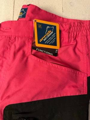 Arrak Outdoor rosa och svarta täckbyxor  - Känns absolut inte inte som denna storlek då jag själv nu mer är stl 38: a. Snygga täckbyxor från Arrak Outdoor i en cool rosa och svart färgkombination. Byxorna har flera praktiska fickor, dragkedjor längs benen och förstärkta partier på knän och bak. Perfekta för utomhusaktiviteter och har en normal passform med elastisk midja. Aldrig använd.