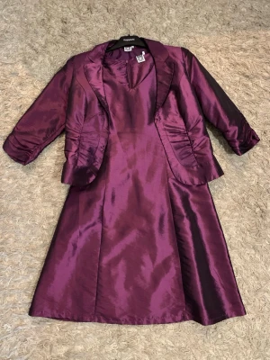 Lila långklänning med matchande kavaj - Säljer en elegant lila långklänning från KappAhl med tillhörande kavaj. Klänningen är ärmlös med v-ringning och har en glansig satinliknande yta. Kavajen har trekvartsärm, snygga rynkdetaljer och klassisk krage. Perfekt för dig som vill sticka ut med en sofistikerad look.