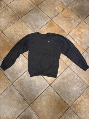 Mörkgrå sweatshirt från Champion - Mörkgrå sweatshirt från Champion med klassisk logga broderad på bröstet. Tröjan har rund hals, långa ärmar och ribbade muddar vid ärmslut och nederkant. Perfekt för chill dagar och enkel att matcha med jeans eller joggers. Storleken står inte på tröjan, men är ungefär en M (kvinna).