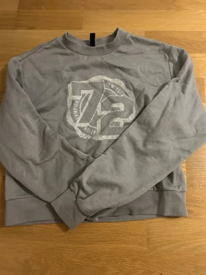 Grå sweatshirt  - Säljer en grå sweatshirt från H&M Divided i storlek XXS. Tröjan har ett stort tryck med siffran 72 och texten 'New York' framtill. Rund hals, ribbade muddar vid ärmslut och nederkant. Perfekt för en chill och avslappnad stil.