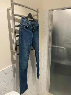 Blå jeans från Nudie Jeans - Säljer ett par klassiska blå jeans från Nudie Jeans med snygga orangea sömmar och broderad detalj på bakfickorna. Perfekt för dig som gillar stilrena och tidlösa jeans. Storlek bredd 30 och längd 32 