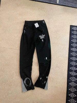 Svarta mjukisbyxor Gallery Dept. - Säljer ett par svarta mjukisbyxor från Gallery Dept. med vitt tryck på ena benet och coola färgstänk i vitt och grönt. Byxorna har resår i midjan med snörning och gråa muddar vid bensluten. Perfekta för en avslappnad och trendig look.