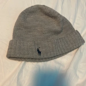 Grå stickad mössa från Polo Ralph Lauren - Snygg grå mössa från Polo Ralph Lauren med klassisk ribbad kant och broderad logga framtill. Stickad i mjuk ull som håller dig varm och ger en clean look. Perfekt för dig som gillar stilrena accessoarer med premiumkänsla.