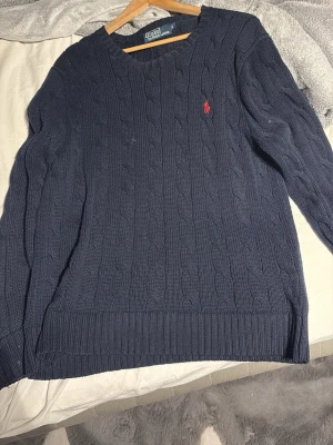 Mörkblå stickad tröja Polo Ralph Lauren - Säljer en mörkblå kabelstickad tröja från Polo Ralph Lauren i storlek S. Tröjan har rund halsringning, långa ärmar och den klassiska röda loggan broderad på bröstet. Perfekt för dig som gillar stilren och tidlös design.