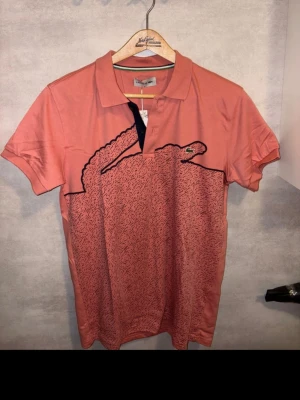 Korallfärgad pikétröja från Lacoste - Säljer en korallfärgad pikétröja från Lacoste med stor grafisk krokodil på framsidan. Tröjan har klassisk krage, korta ärmar och knappar vid halsen. Materialet är mjuk bomull och insidan av kragen har en grön och vit rand. Perfekt för dig som gillar statement-plagg.