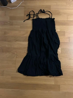 Svart långklänning från Ellos - Svart långklänning från Ellos med smock upptill och knytbara axelband. Klänningen har en luftig och böljande kjol med volangdetaljer, perfekt för varma dagar. Materialet känns mjukt och lätt, troligen bomull. Enkel och stilren design.