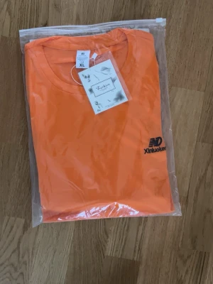 Orange t-shirt från Xinluokent XL - Snygg orange t-shirt från Xinluokent i storlek XL. Klassisk rund halsringning och korta ärmar. T-shirten har en enkel design med en liten svart logga på bröstet. Tillverkad i mjuk bomull som känns skön mot huden.