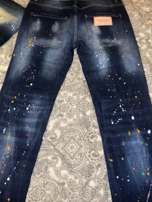 Mörkblå jeans med färgstänk från Dsquared2 - Säljer ett par mörkblå jeans från Dsquared2 med coola färgstänk i orange, vitt, grönt och rött. Jeansen har slitna detaljer och hål på framsidan för en edgy vibe. Klassisk femficksmodell med Dsquared2-lapp bak. Perfekt för dig som gillar streetstyle.