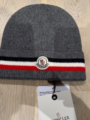 Grå Moncler mössa med ränder - Säljer en snygg grå mössa från Moncler med ribbad struktur och klassiska Moncler-loggan framtill. Mössan har grå, vita och röda ränder längs kanten som ger en sportig vibe. Perfekt för kalla dagar och streetstyle.