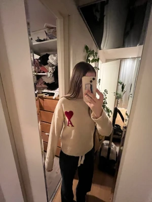 Beige stickad tröja från Ami Paris - Säljer en beige stickad tröja från Ami Paris med röd hjärtlogga och bokstaven A på bröstet. Tröjan har rund hals, långa ärmar och ribbade muddar. Perfekt för dig som vill ha en mysig och snygg statementtröja.