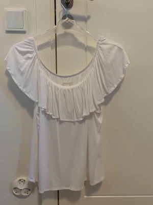 Vit topp med volang från H&M - Säljer en vit topp från H&M i storlek M. Toppe  har bred volang runt halsringningen och är offshoulder med omlott-design. Tillverkad i mjuk och lätt viskos som ger ett luftigt intryck. Perfekt för varma dagar och sommarhäng. Aldrig använd dock tvättad en gång 