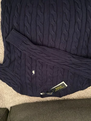 Marinblå kabelstickad tröja Polo Ralph Lauren - Snygg marinblå kabelstickad tröja från Polo Ralph Lauren med klassisk broderad logga på bröstet. Tröjan har långärmade ribbade muddar och rund hals. Perfekt för dig som gillar stilrena och tidlösa plagg med premiumkänsla.