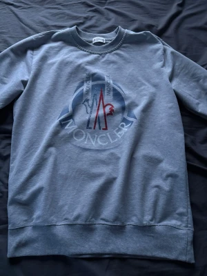 Grå sweatshirt från Moncler, M - Säljer en grå sweatshirt från Moncler i storlek M. Tröjan har ett stort tryckt Moncler-logo i blått, rött och vitt på bröstet. Rund hals, ribbade muddar vid ärmslut och nederkant. Perfekt för dig som gillar stilrena märkesplagg. Pris kan diskuteras vid snabb affär!