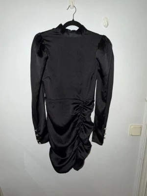 Svart satin klänning med puffärm - Snygg svart kortklänning i satin med puffärmar och markerade axlar. Klänningen har lång ärm med dekorativa knappar vid ärmslut och cool rynkning längs kjolen som ger en unik look. Perfekt för dig som vill sticka ut.