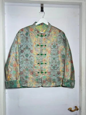 Silkesjacka i 100 % siden – konstnärligt mönster  - Elegant jacka i 100 % siden med ett rikt, måleriskt blommönster i varma gröna och gyllene toner. Tyget är mjukt, lätt och följsamt med en naturlig lyster som ger plagget ett exklusivt uttryck.  Avslappnad passform som faller fint på kroppen. De dekorativa knäppningarna framtill ger en subtil orientalisk känsla och gör jackan både personlig och tidlös.  🧵 Material 	•	100 % siden
