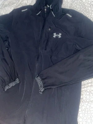 Svart vindjacka från Under Armour - Svart vindjacka från Under Armour med huva och dragkedja framtill. Jackan har lång ärm med logotypdetaljer på mudden och bröstet. Perfekt för träning utomhus och har en sportig look med snygga reflexdetaljer.