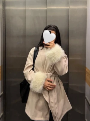 Beige kappa med pälsdetaljer - Säljer en beige kappa med fluffiga vita pälsdetaljer vid ärmsluten och ett bälte i midjan. Jackan har en omlottstil och ger en riktigt mysig och trendig vibe. Perfekt för dig som vill ha en stilren look med lite extra lyxig känsla.😍❤️