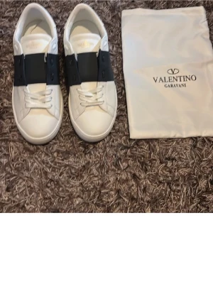 Valentino Garavani vita sneakers - Säljer ett par snygga vita Valentino Garavani sneakers med bred svart rem över vristen och vita snören. Skorna har rund tå, platt sula med mönstrad undersida och kommer med original dustbag. Tillverkade i skinn för en lyxig känsla.
