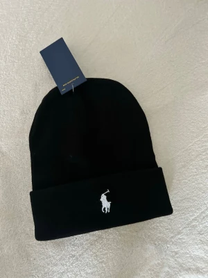 Svart mössa från Polo Ralph Lauren - Svart mössa från Polo Ralph Lauren med klassisk broderad logga i vitt framtill. Mössan har uppvikt kant och är tillverkad i mjukt material som håller dig varm. Perfekt accessoar för kyliga dagar och enkel att matcha med din stil.