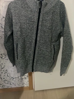 Grå stickad kofta med fleecefoder - Säljer en gråmelerad stickad kofta med dragkedja framtill och två fickor. Insidan är fodrad med mjuk fleece i rutigt mönster, vilket gör den extra skön och varm. Perfekt för kyliga dagar och har en klassisk, enkel look.