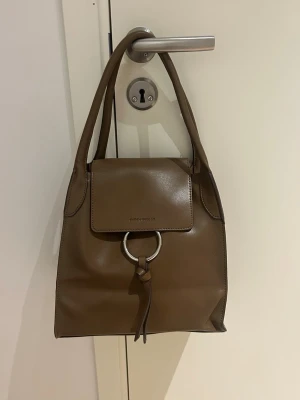 Brun axelväska från Carin Wester - Snygg Taupé/ brun axelväska från Carin Wester i en stilren design med både släta och mocka-detaljer. Väskan har ett stort metallring-spänne framtill och praktiska handtag. Perfekt storlek för att få plats med allt du behöver och har en mjuk insida i mocka. Storlek 27x27x15.