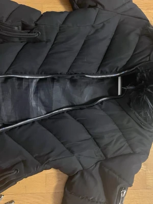 Svart pufferjacka med fuskpäls - Säljer en svart pufferjacka från Bik Bok med fuskpäls på huvan. Jackan har dragkedja framtill, snörning i midjan och flera fickor med dragkedja. Perfekt för kalla dagar och riktigt snygg till streetwear-stilen.