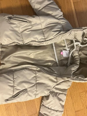 Beige pufferjacka från Vero Moda - Säljer en beige pufferjacka från Vero Moda med huva och två sidofickor med dragkedja. Jackan har quiltad design och är tillverkad i syntetmaterial, perfekt för kyliga dagar. Stängs med dragkedja framtill och har en loose passform.