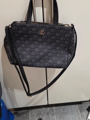 Svart handväska från Lycke Oslo - Snygg svart handväska från Lycke Oslo med justerbar axelrem och guldfärgade detaljer. Väskan har ett mönster med logotypen tryckt över hela ytan och flera praktiska fack med dragkedja. Materialet är skinnimitation och designen är stilren med både handtag och axelrem.