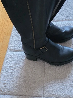 Svarta boots med grov sula och dragkedja - Säljer ett par svarta boots i skinn med grov platt sula och låg klack. De har dragkedja på sidan och ett snyggt spänne över foten. Insidan är fodrad med mjukt material för extra komfort. Perfekta för höst och vinter när du vill ha både stil och funktion.