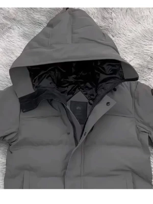 Grå dunjacka från Canada Goose - Snygg grå dunjacka från Canada Goose med huva och svart logotyp-patch på ärmen. Jackan har dragkedja och knappar framtill, två sidofickor och quiltad design. Perfekt för kalla dagar och har en clean, modern look. Det finns inne i M men även L 