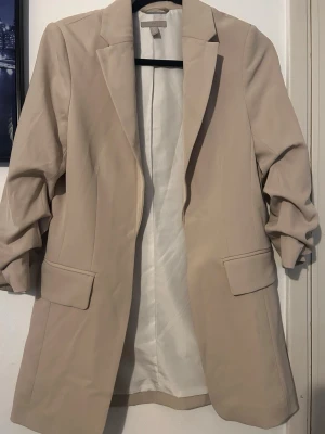 Beige kavaj från H&M, XS.  - Snygg beige kavaj från H&M i klassisk modell med krage och två fickor framtill. Lätt figursydd och med långa ärmar, perfekt för en clean och stilren look. Materialet känns mjukt och har en lätt glansig finish. Passar dig som gillar minimalistisk stil.