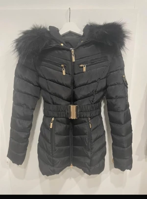 Svart pufferjacka med päls från Roccobarocco - Svart pufferjacka från Roccobarocco med fluffig päls på huvan, guldfärgade dragkedjor och detaljer, samt bälte i midjan. Jackan har flera fickor med dragkedja och knapp, quiltad design och lång ärm. Fodret har leopardmönster för extra edge. fuskpäls